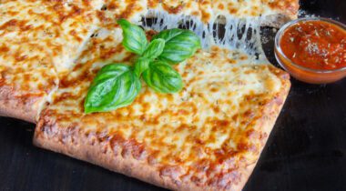 slice of square pizza pulled away from whole pizza. basil on top of pizza slice and cup of sauce on black tabletop background